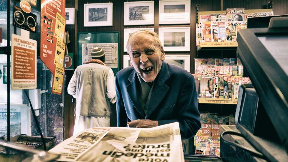 An older man smiling as he holds up a newspaper in a shop
