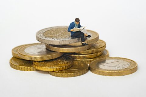 Small figurine sitting on a stack of coins reading a newspaper