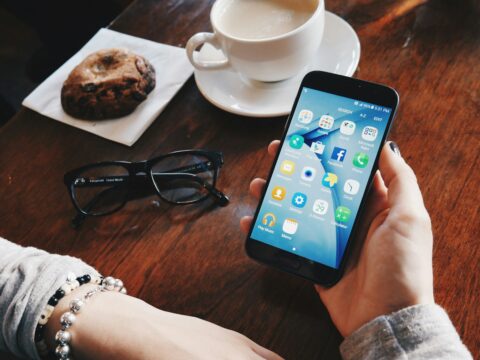 Hand holding a smartphone at a table with a coffee cup and glasses